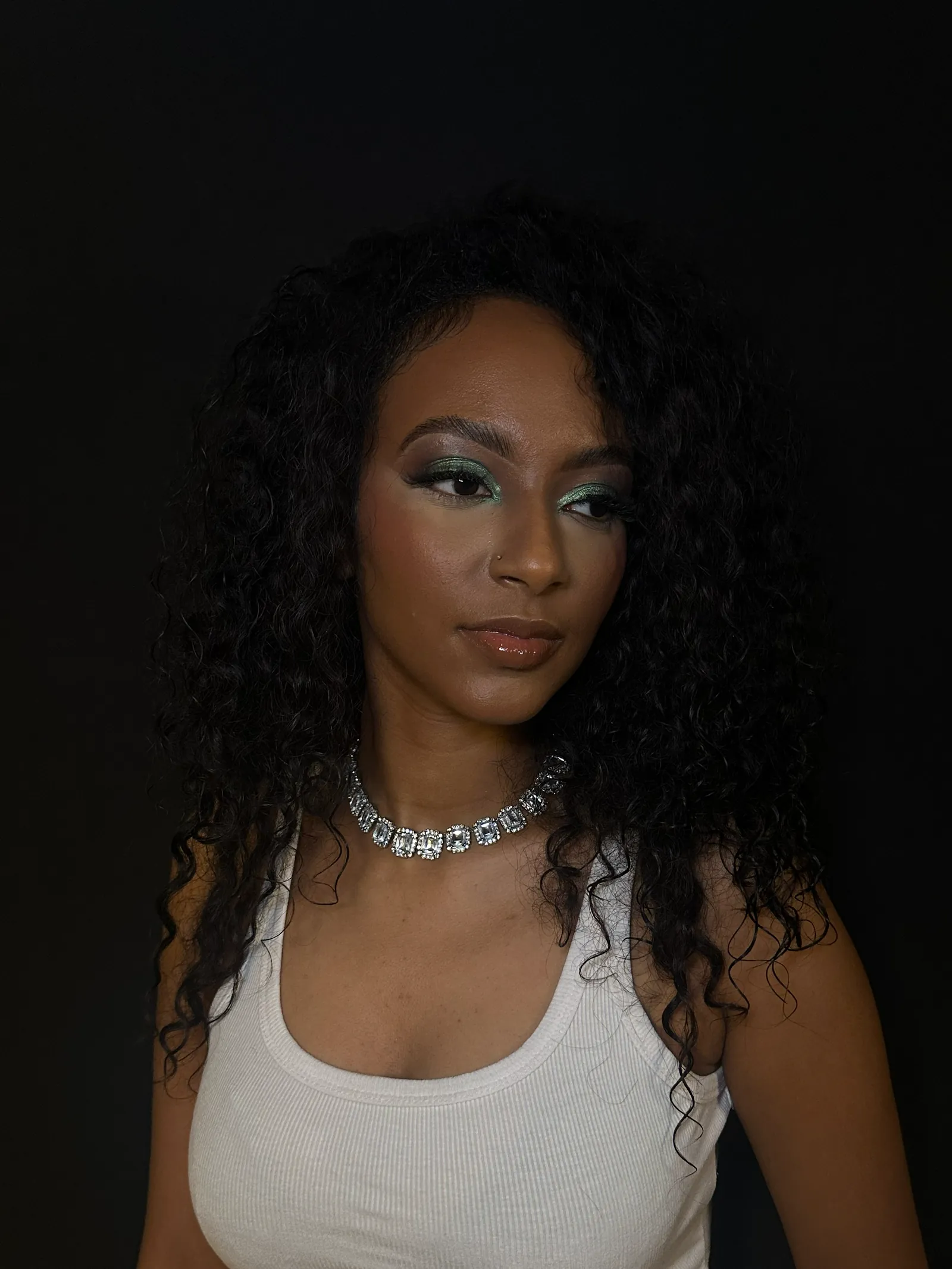 Teal eyeshadow, glossy nude lips and a crystal necklace against a dark background
