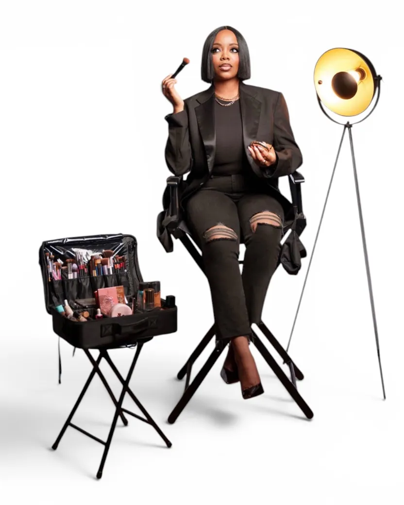 Keren in a black blazer and ripped black jeans, seated in a director's chair with a brush raised, beside her open makeup case and a studio light