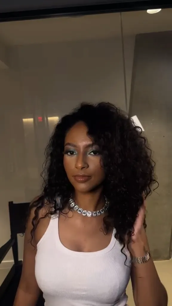 Woman with teal shimmer eyeshadow and a crystal necklace posing in a studio, hands in her curls