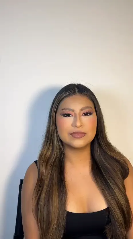 Woman with long brown hair, pink eyeshadow and blush, turning towards the camera
